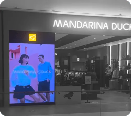 Entrance to Mandarina Duck store with illuminated sign and large digital display showing two models wearing blue sweatshirts.