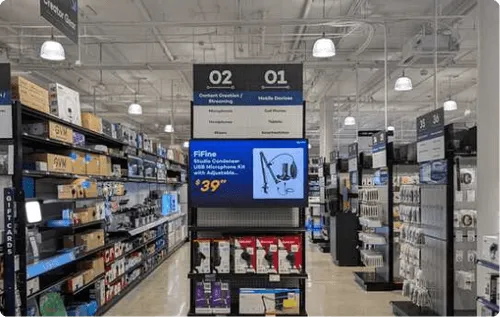Electronics aisle in a store with shelves displaying headphones, microphones, and other audio accessories under signs labeled 'Content Creators/Streaming' and 'Mobile Devices'.