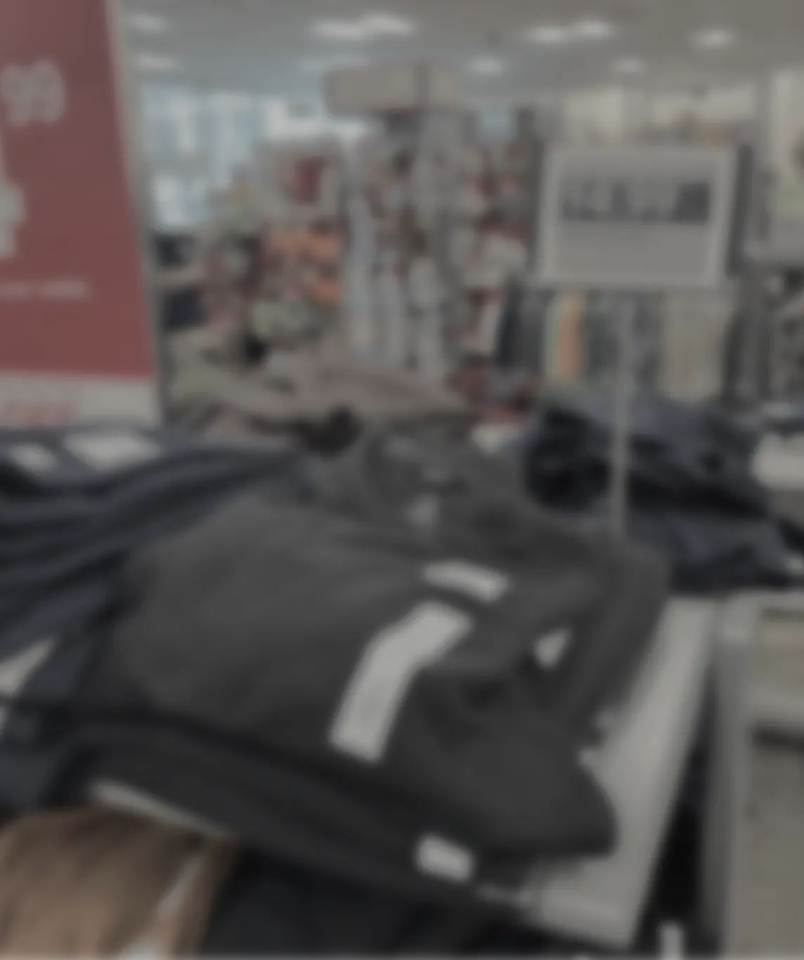 Blurred image of folded clothing items on display tables inside a retail store with shelves and product racks in the background